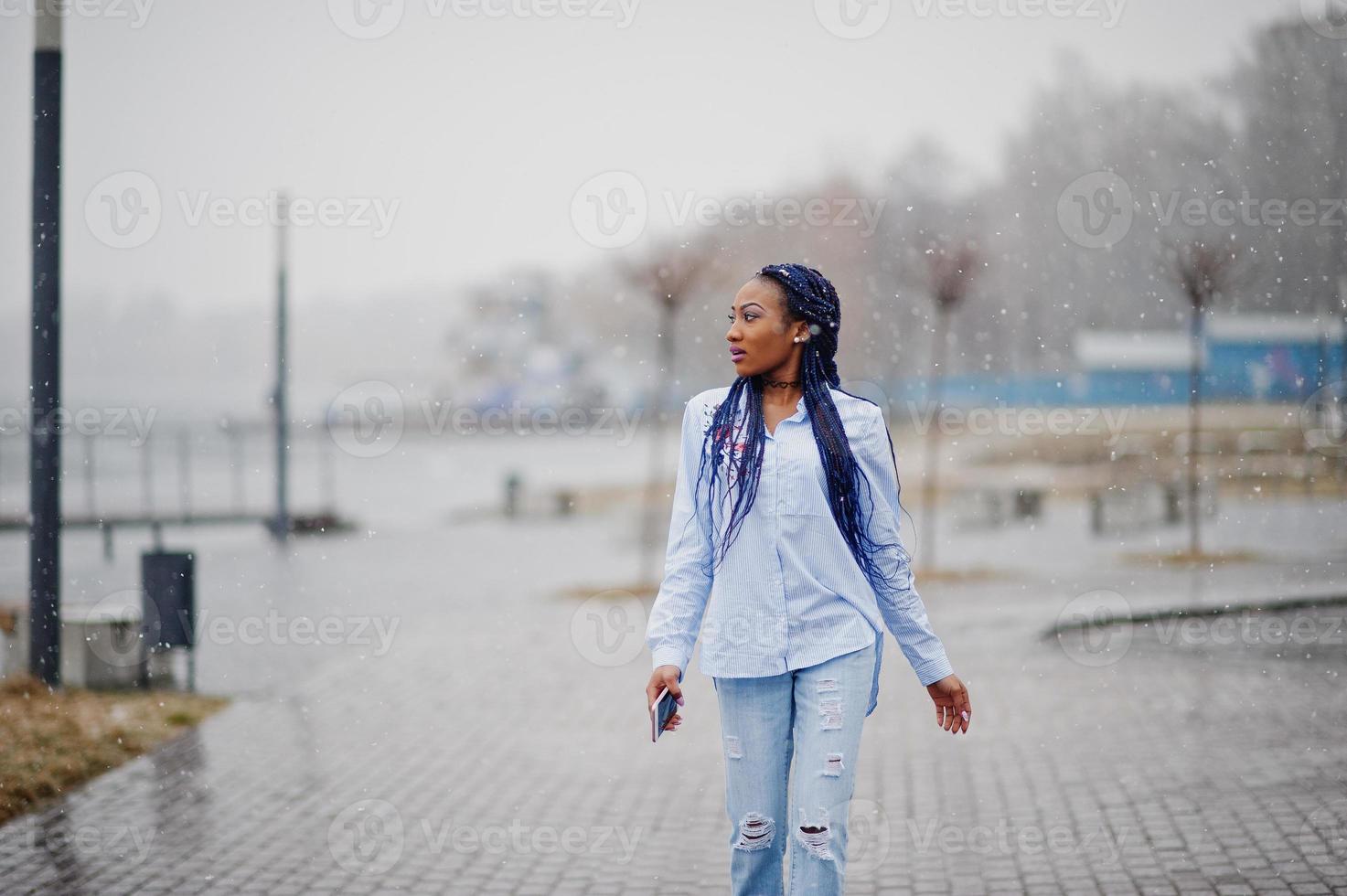 elegante chica afroamericana con rastas sosteniendo el teléfono móvil a mano, al aire libre con clima nevado. foto