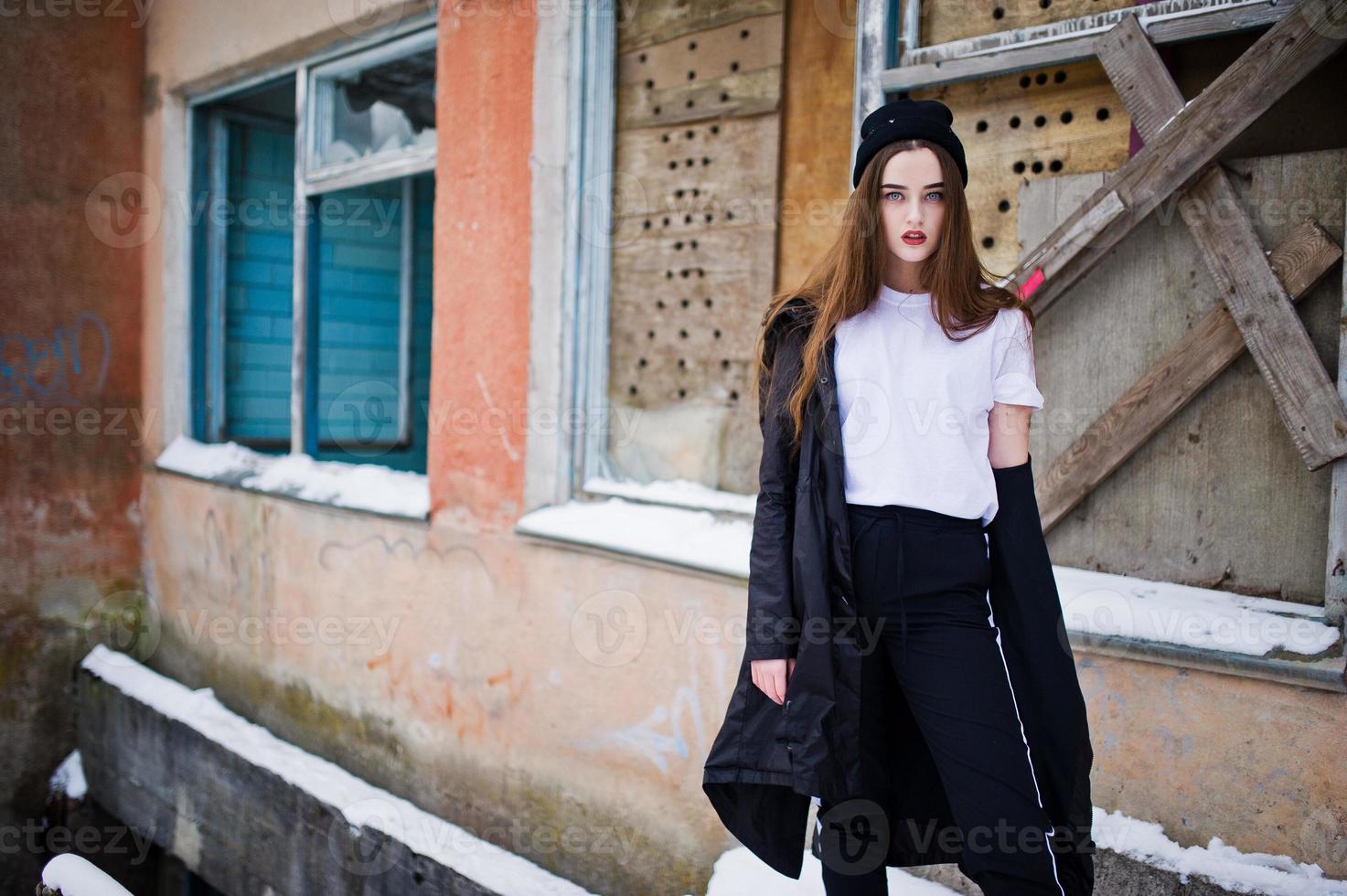 Fashionable long legs brunette model in long black cloak posed outdoor at winter day against old grunge wall with broken windows. photo