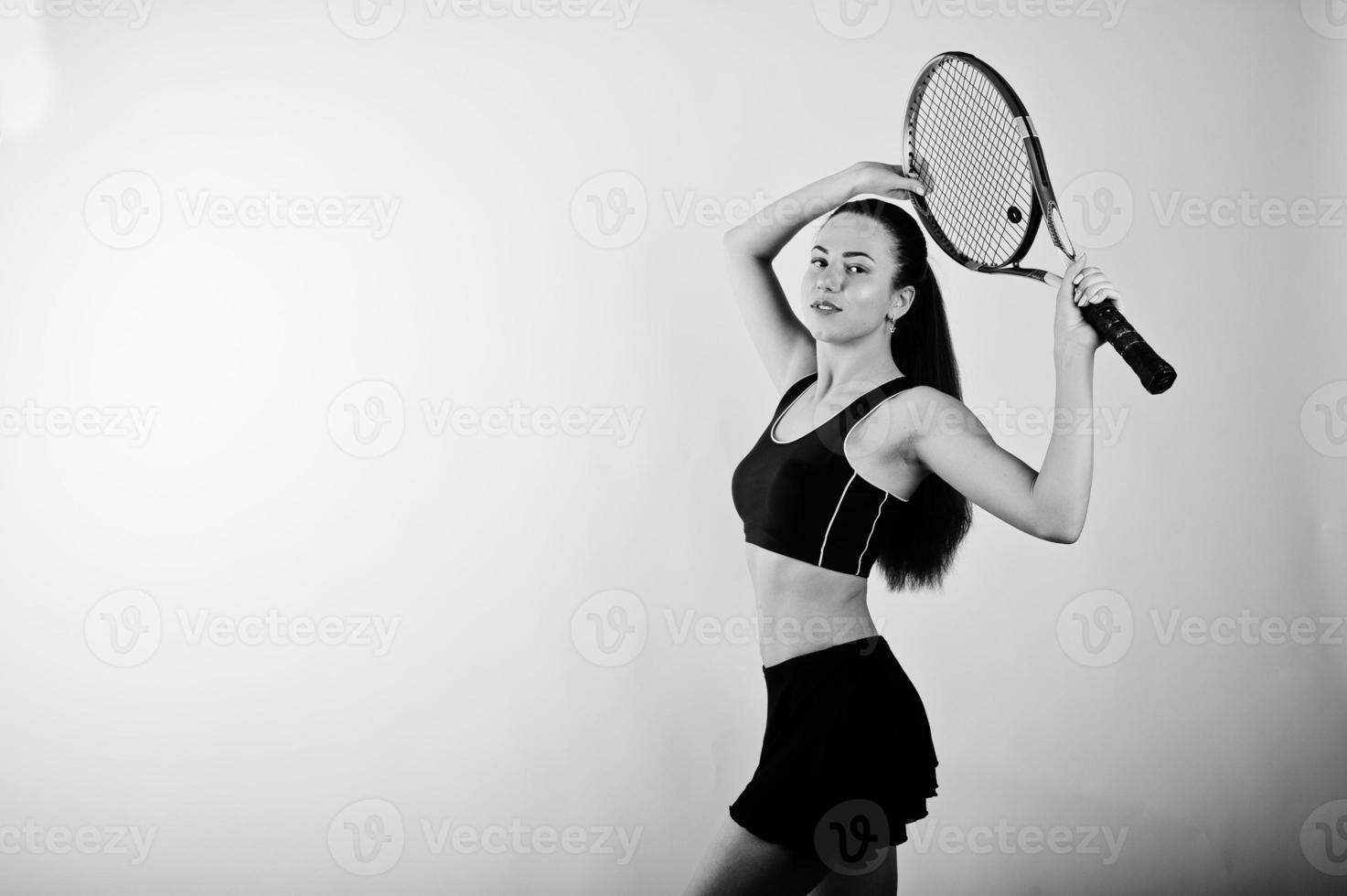 Black and white portrait of beautiful young woman player in sports clothes holding tennis racket while standing against white background. photo