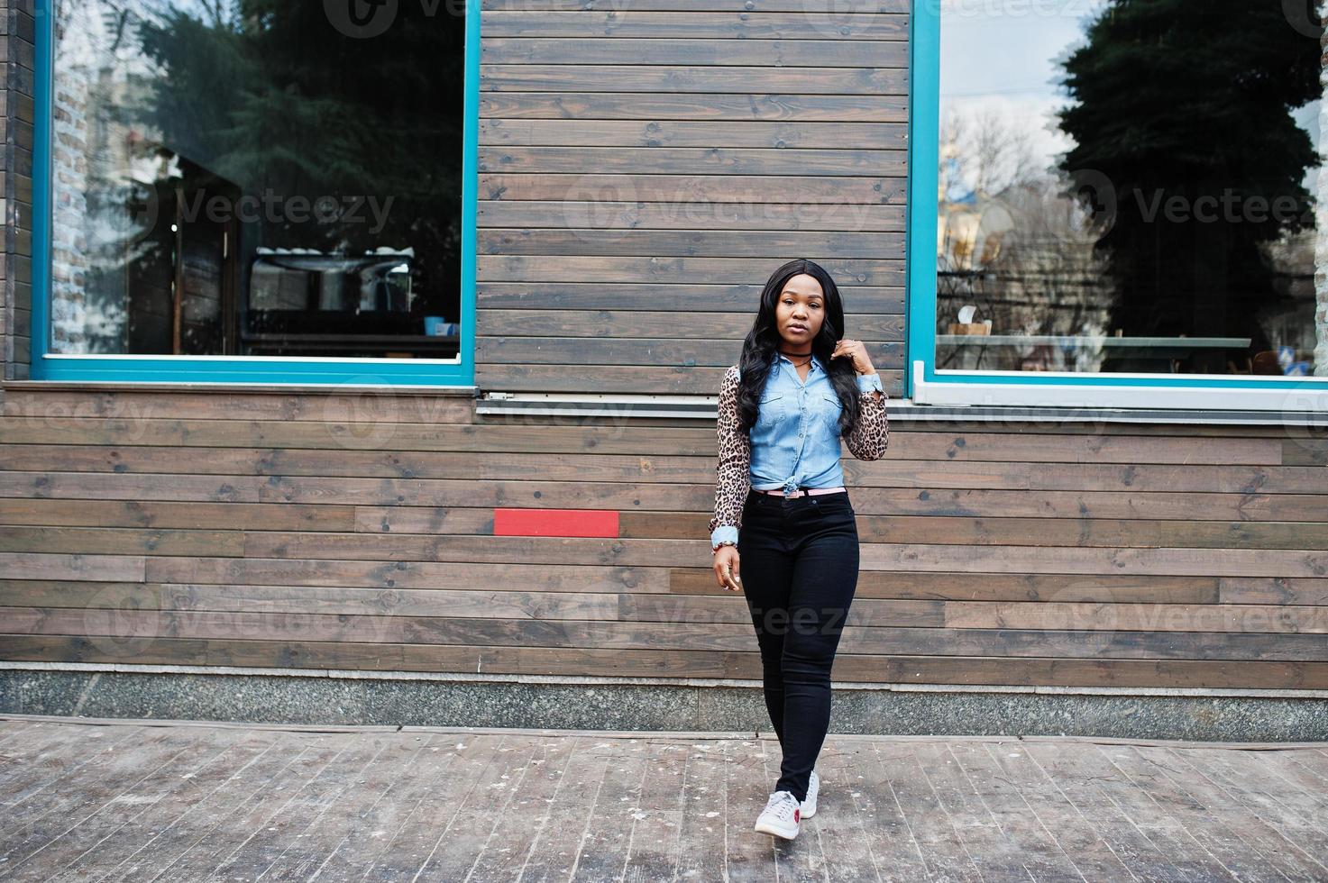 chica afroamericana hipster con camisa de jeans con mangas de leopardo posando en la calle contra una casa de madera con ventanas. foto