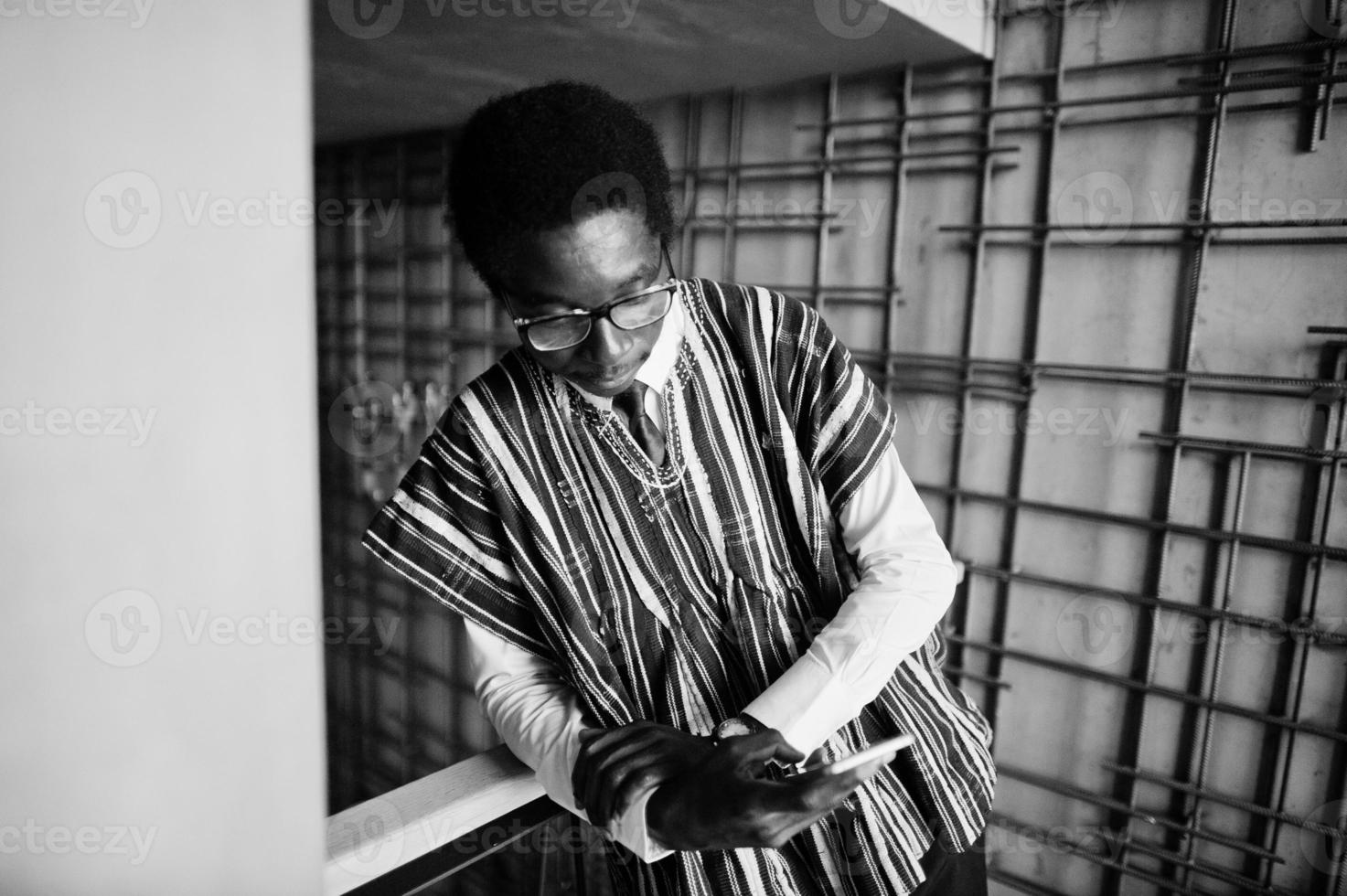 African businessman in traditional clothes and glasses with mobile phone at hand posed at modern building indoor. photo