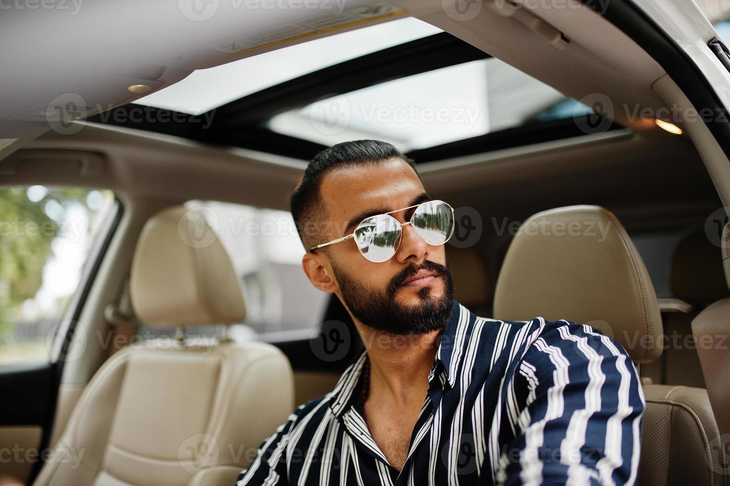 Successful arab man wear in striped shirt and sunglasses pose behind the wheel of  his white suv car. Stylish arabian men in transport. photo