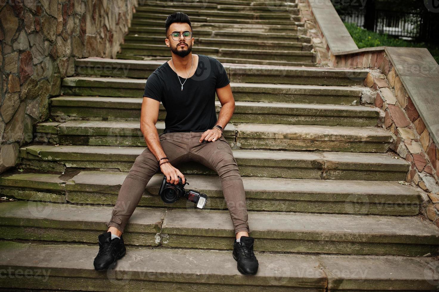 Awesome beautiful tall ararbian beard macho man photographer in glasses and black t-shirt with professional camera at hands. photo