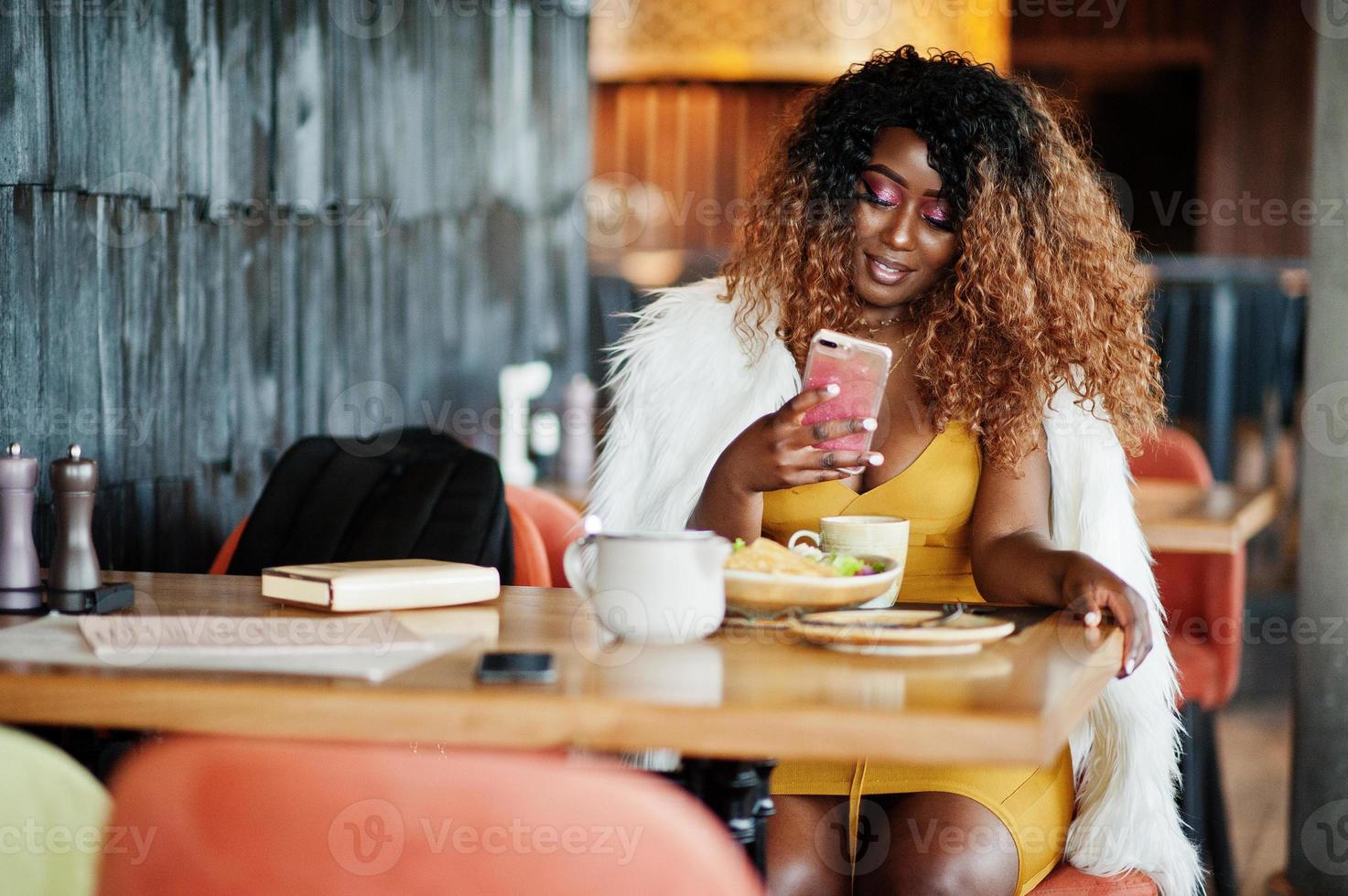 mujer afroamericana glamurosa vestida de amarillo sentada en la mesa con platos en el restaurante con teléfono móvil a mano. foto