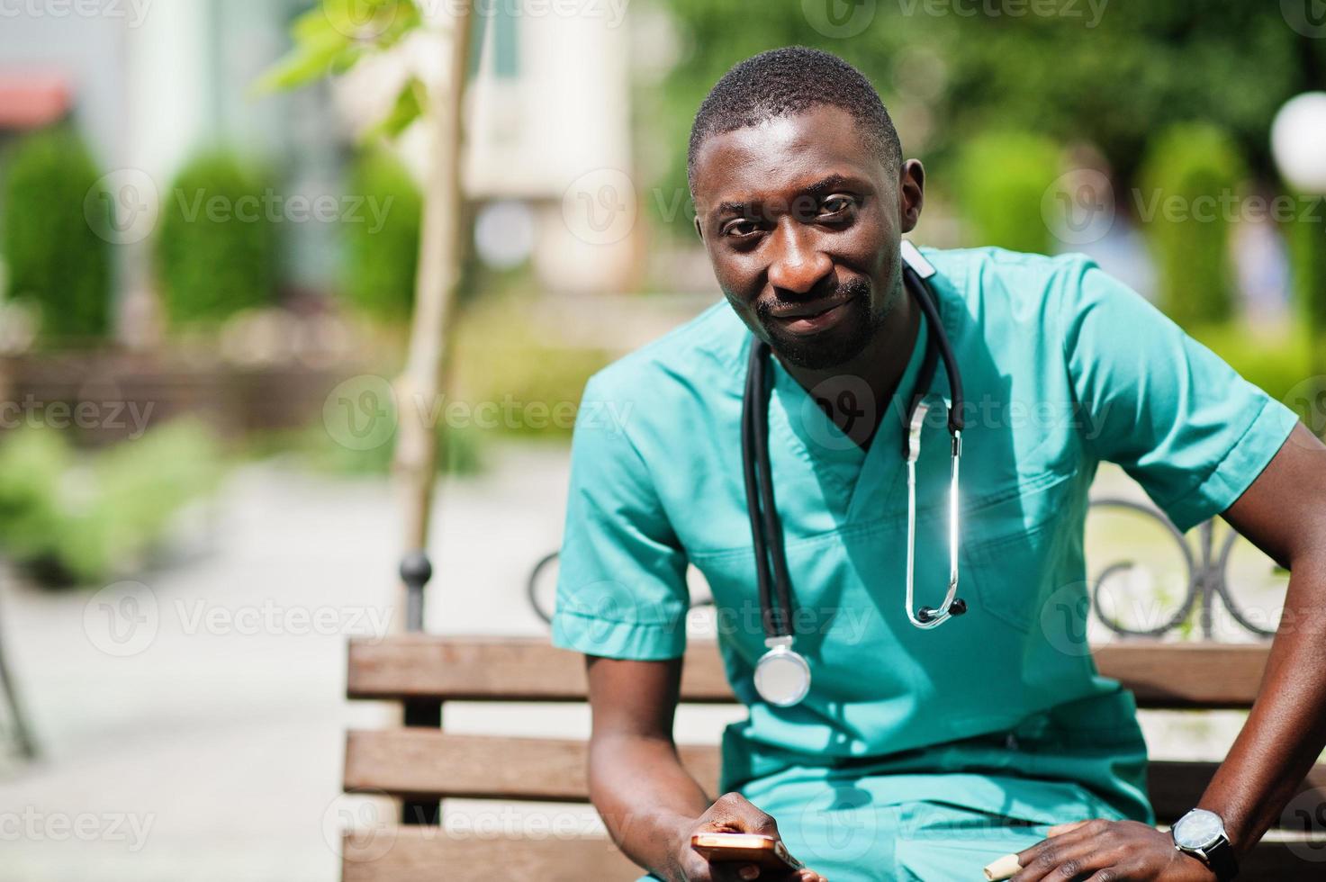 Portrait of African male doctor with stethoscope wearing green coat with mobile phone at hand. photo