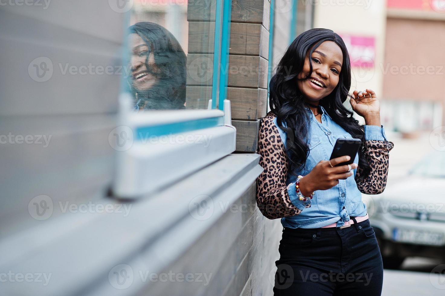 chica afroamericana hipster con camisa de jeans con mangas de leopardo, sostiene el teléfono móvil a mano y posa en la calle contra una casa de madera con ventanas. foto