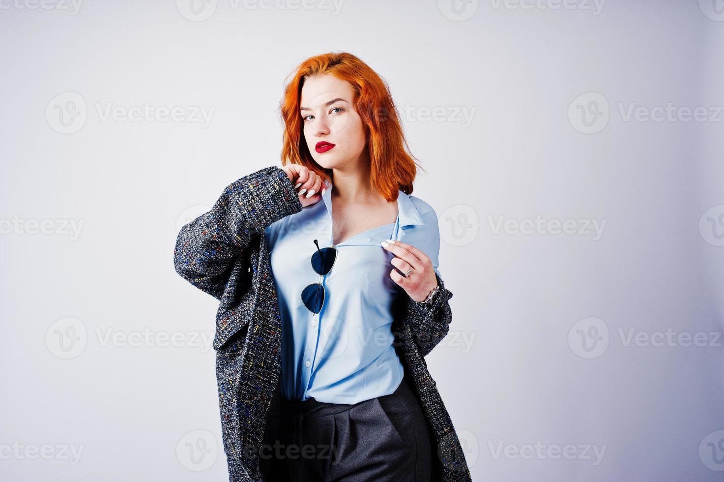 Portrait of a fantastic redheaded girl in blue shirt, grey overcoat posing with sunglasses in the studio. photo