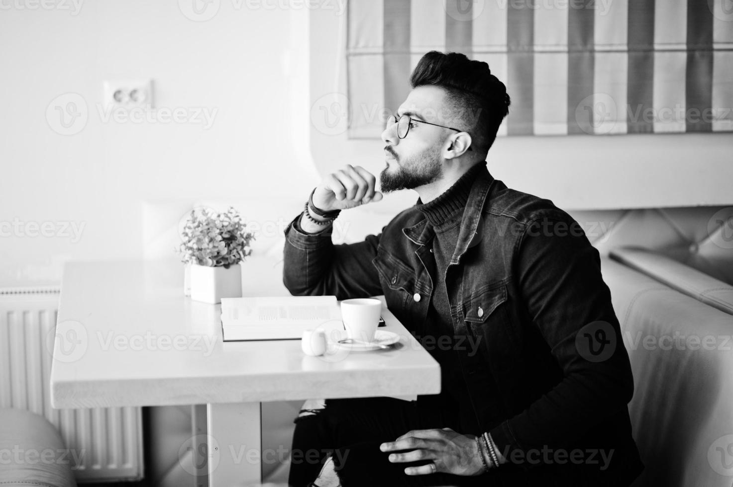 el hombre árabe usa una chaqueta de jeans negros y anteojos sentados en un café, lee un libro y bebe café. chico modelo árabe elegante y de moda. foto