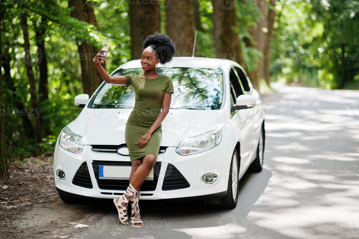 African american woman posed against white car in forest road and looking at mobile phone. photo