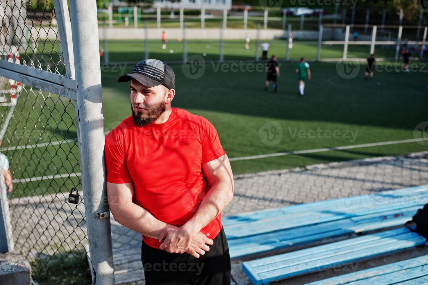 Young brutal bearded muscular man wear on red shirt, shorts and cap at stadium. photo
