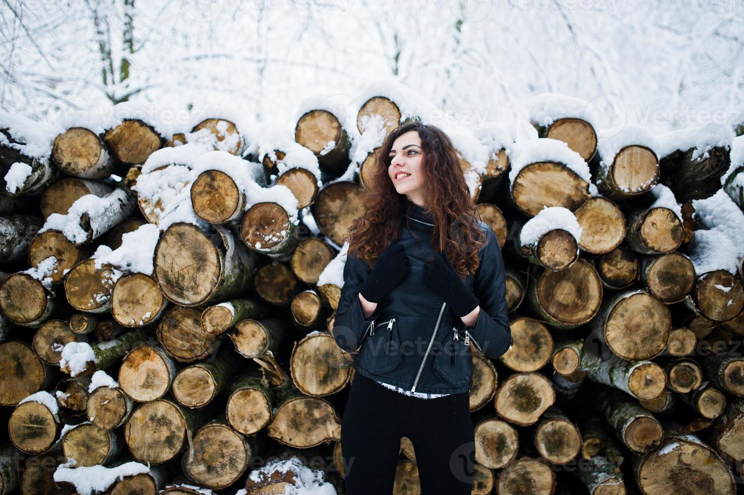 Elegance curly girl in leather jacket at snowy forest park at winter against stumps. photo