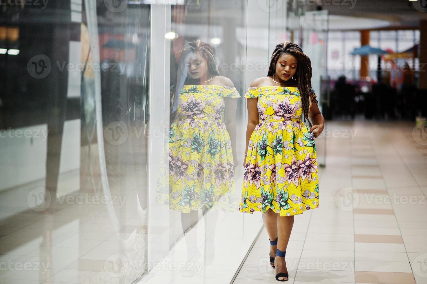 linda chica afroamericana de pequeña altura con rastas, vestida con un vestido amarillo de color, posada en el escaparate de la tienda en el centro comercial. foto