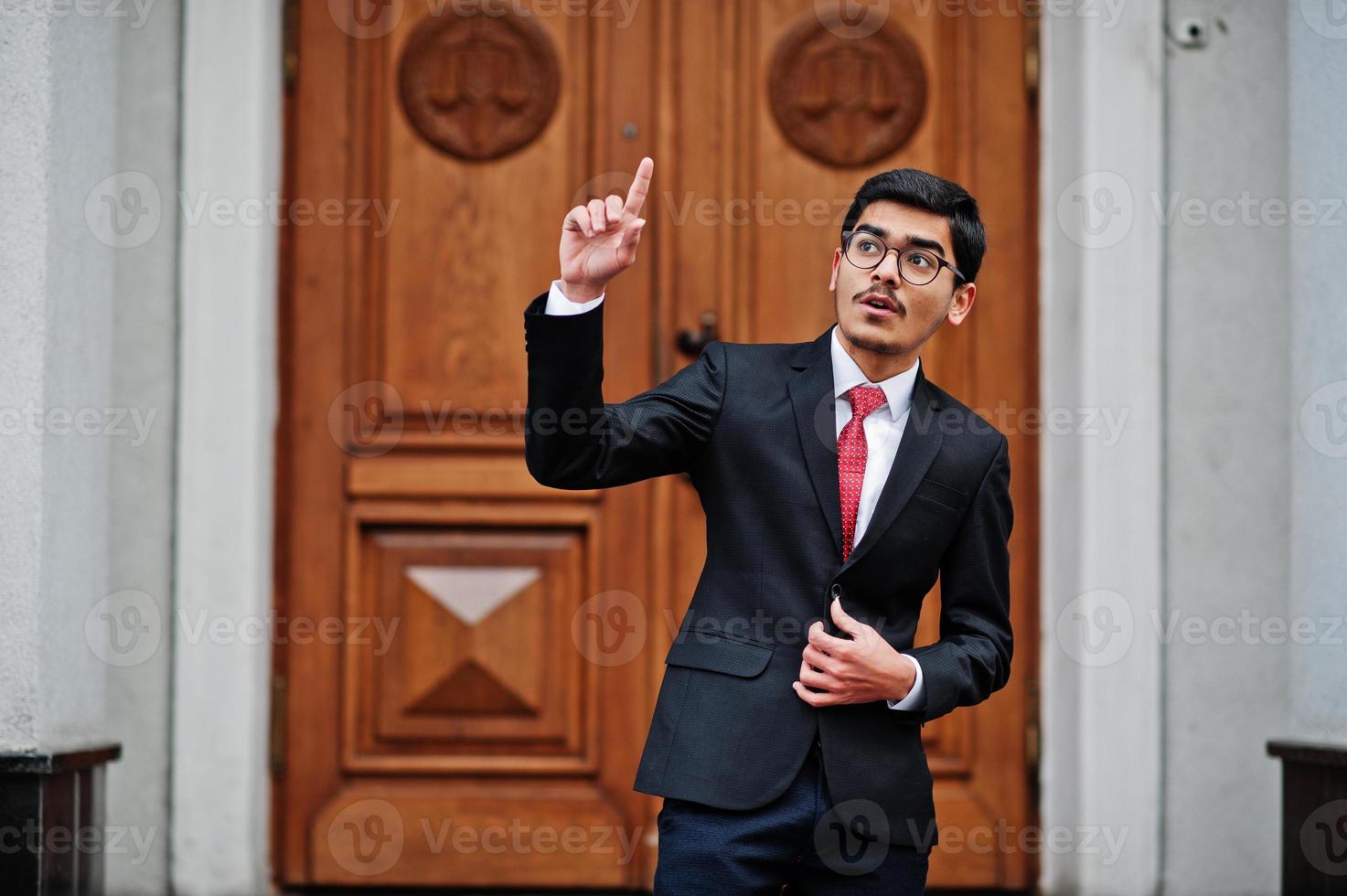 Indian young man at glasses, wear on black suit with red tie posed outdoor against door of building show finger up, he has idea. photo