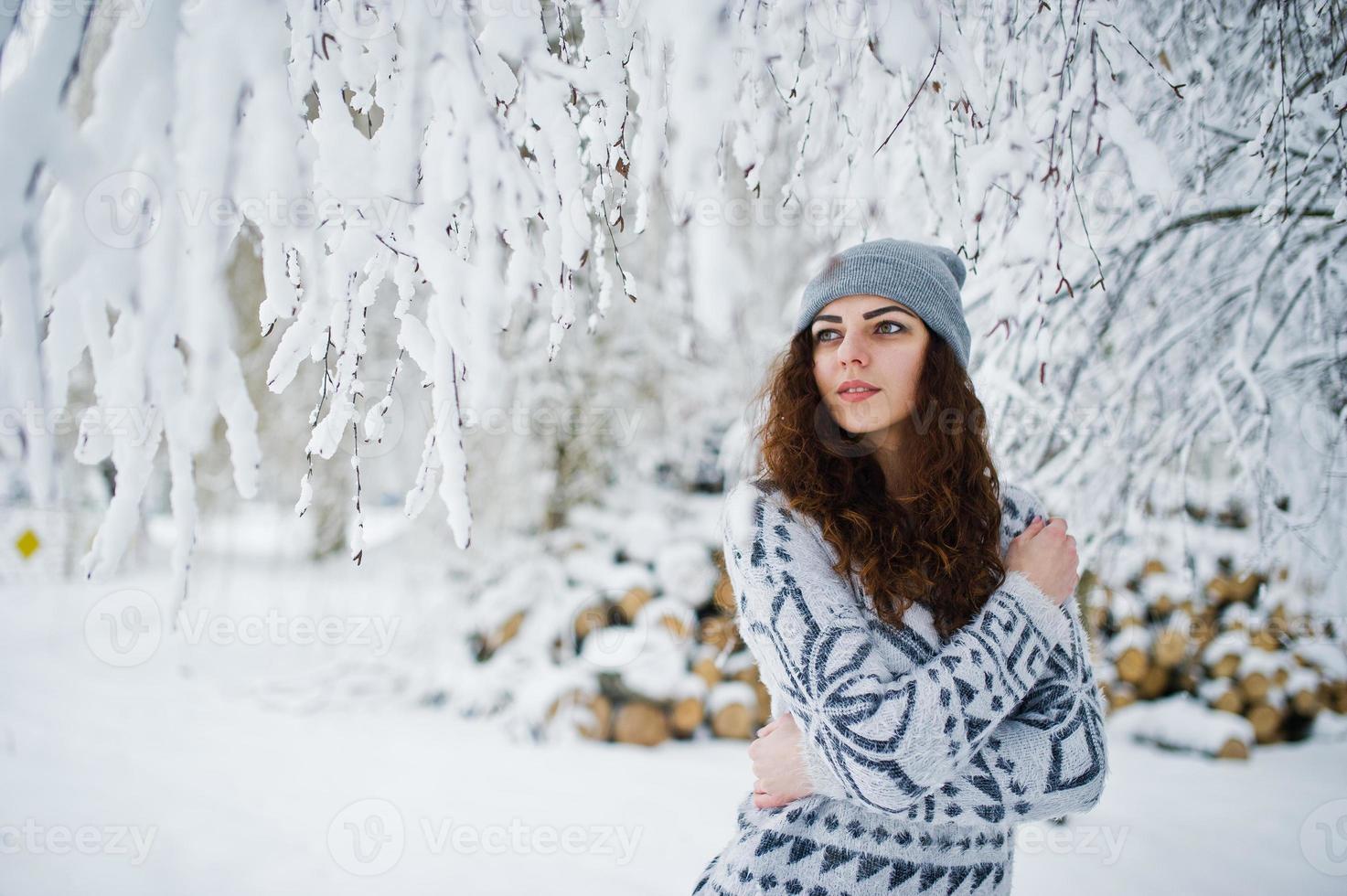 Cute curly girl in sweater and headwear at snowy forest park at winter. photo