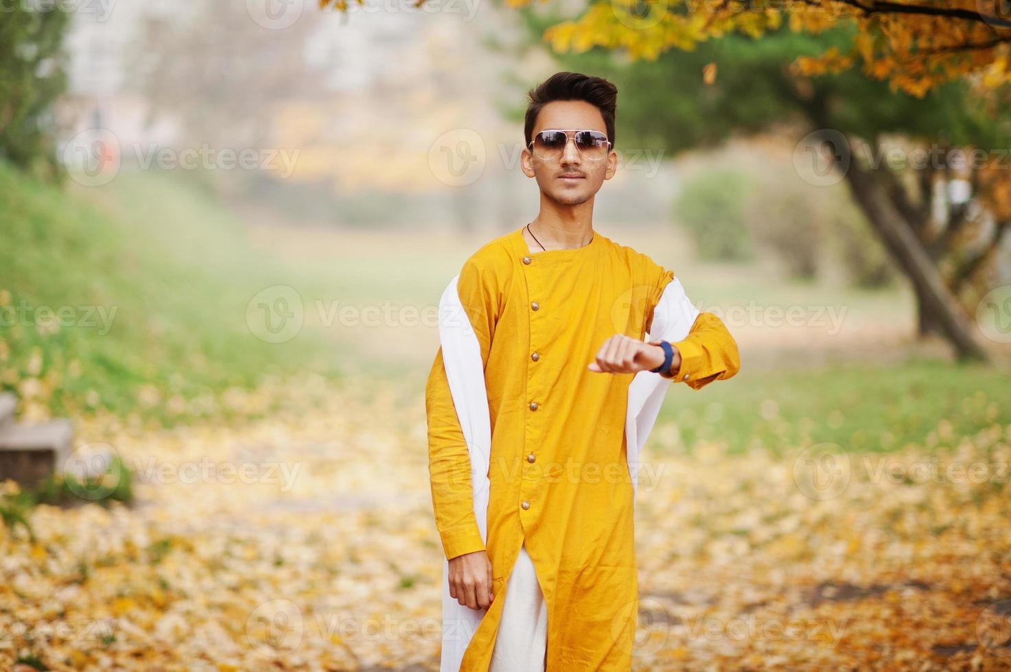 Indian stylish man in yellow traditional clothes with white scarf, sunglasses posed outdoor against autumn leaves tree. photo