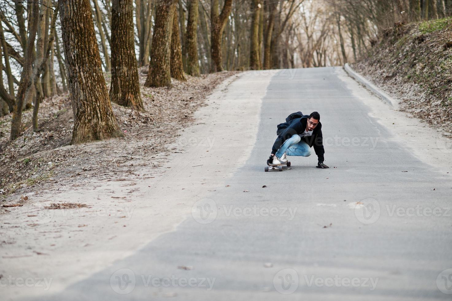 hombre árabe de estilo callejero en anteojos con longboard longboarding por el camino. foto