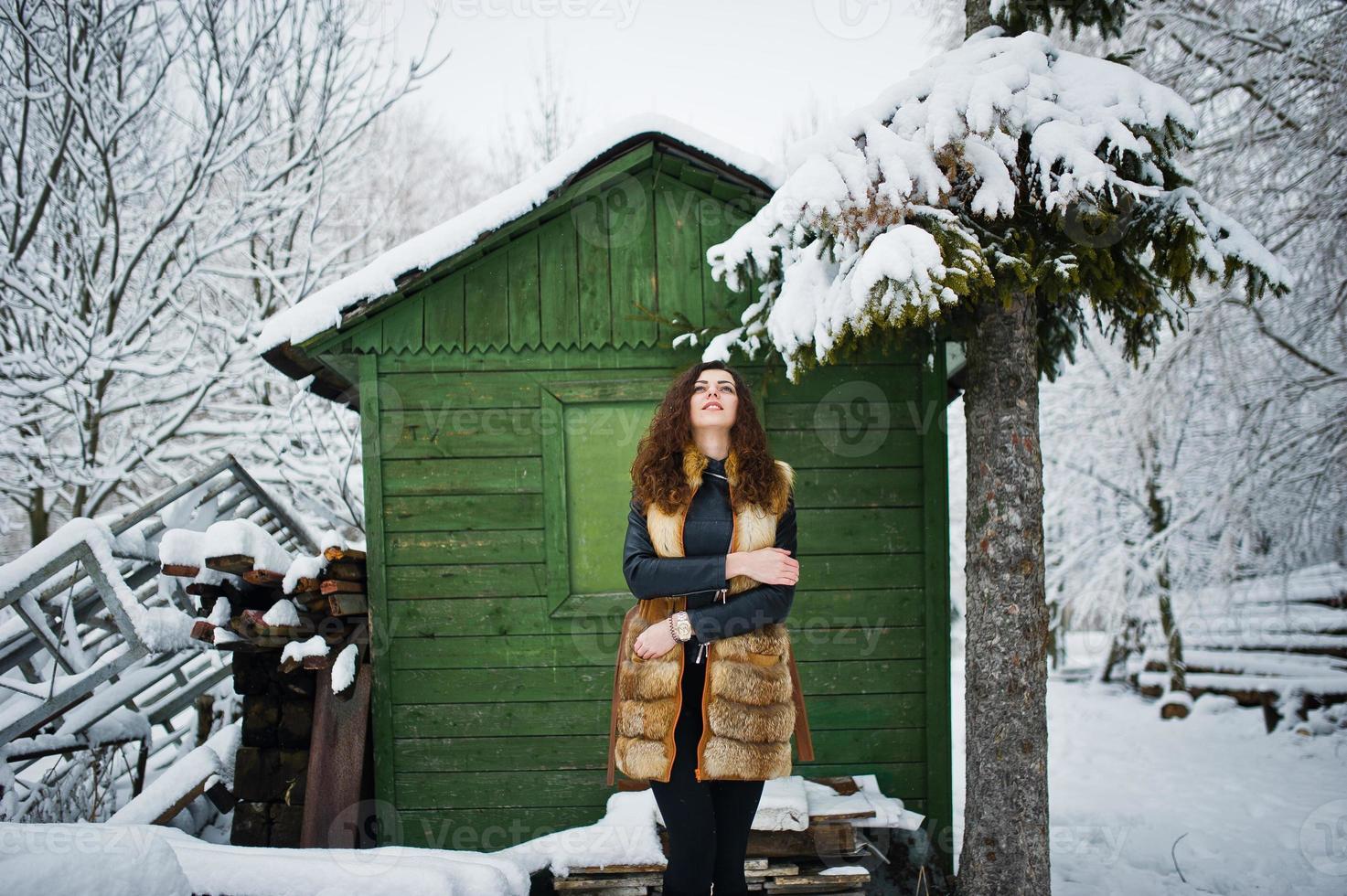 Elegance curly girl in fur coat at snowy forest park agasinst green wooden house at winter. photo