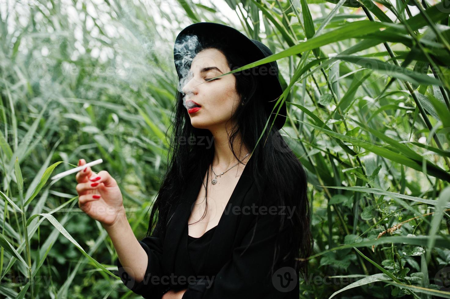 Sensual smoker girl all in black, red lips and hat. Goth dramatic woman smoking on common reed. photo