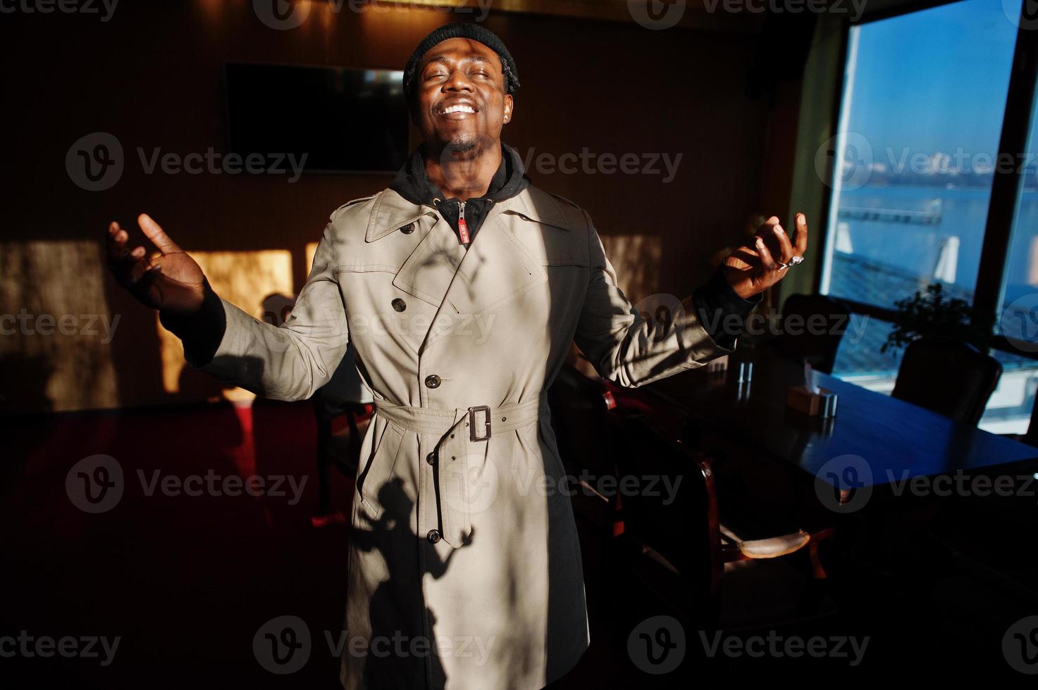 Handsome african american man posing  inside room with sunlight shadows in black hat and beige coat. photo