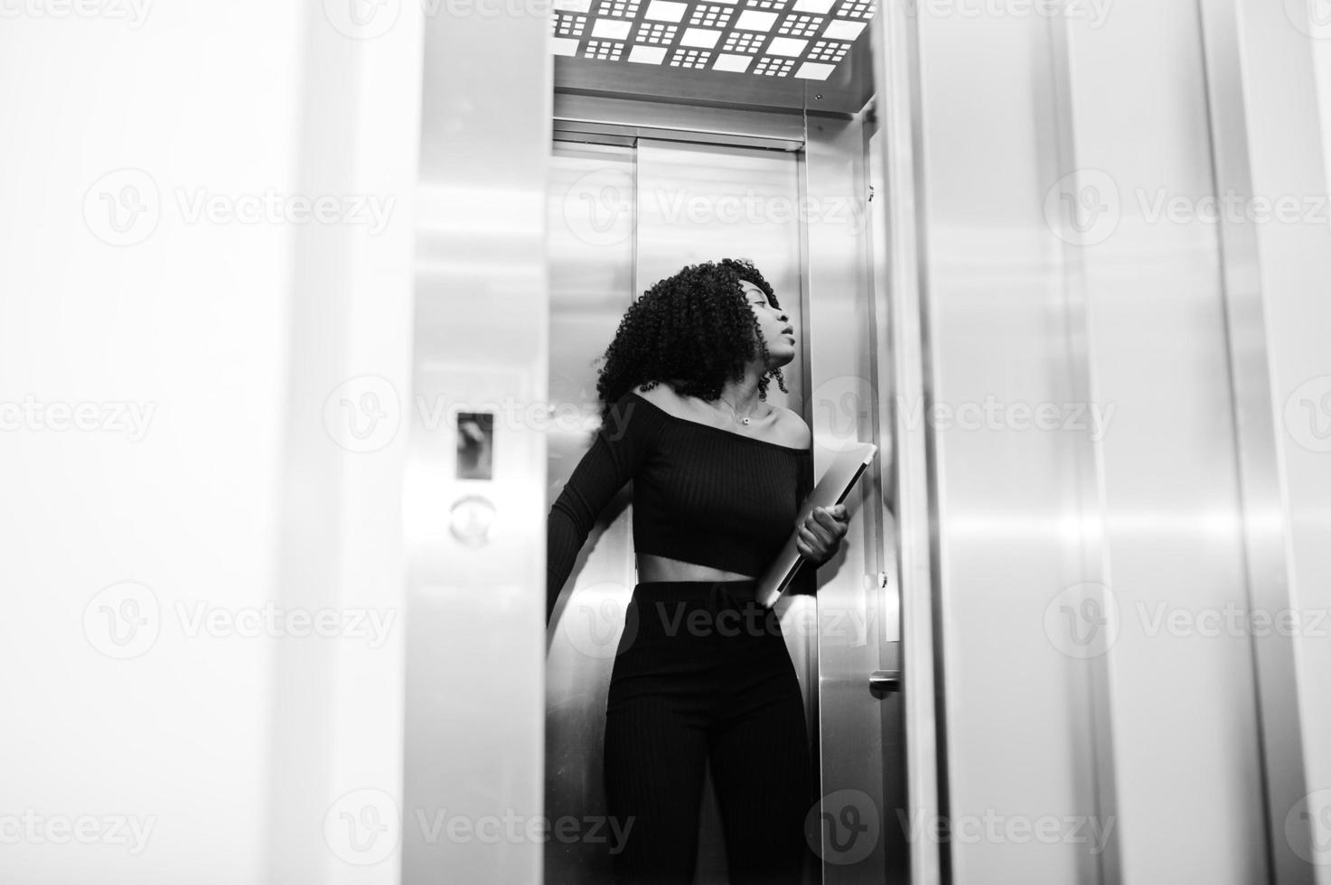 Fashionable young beautiful african american business woman with afro hairstyle wear in elegant black, stand at elevator with phone and laptop at hands. photo