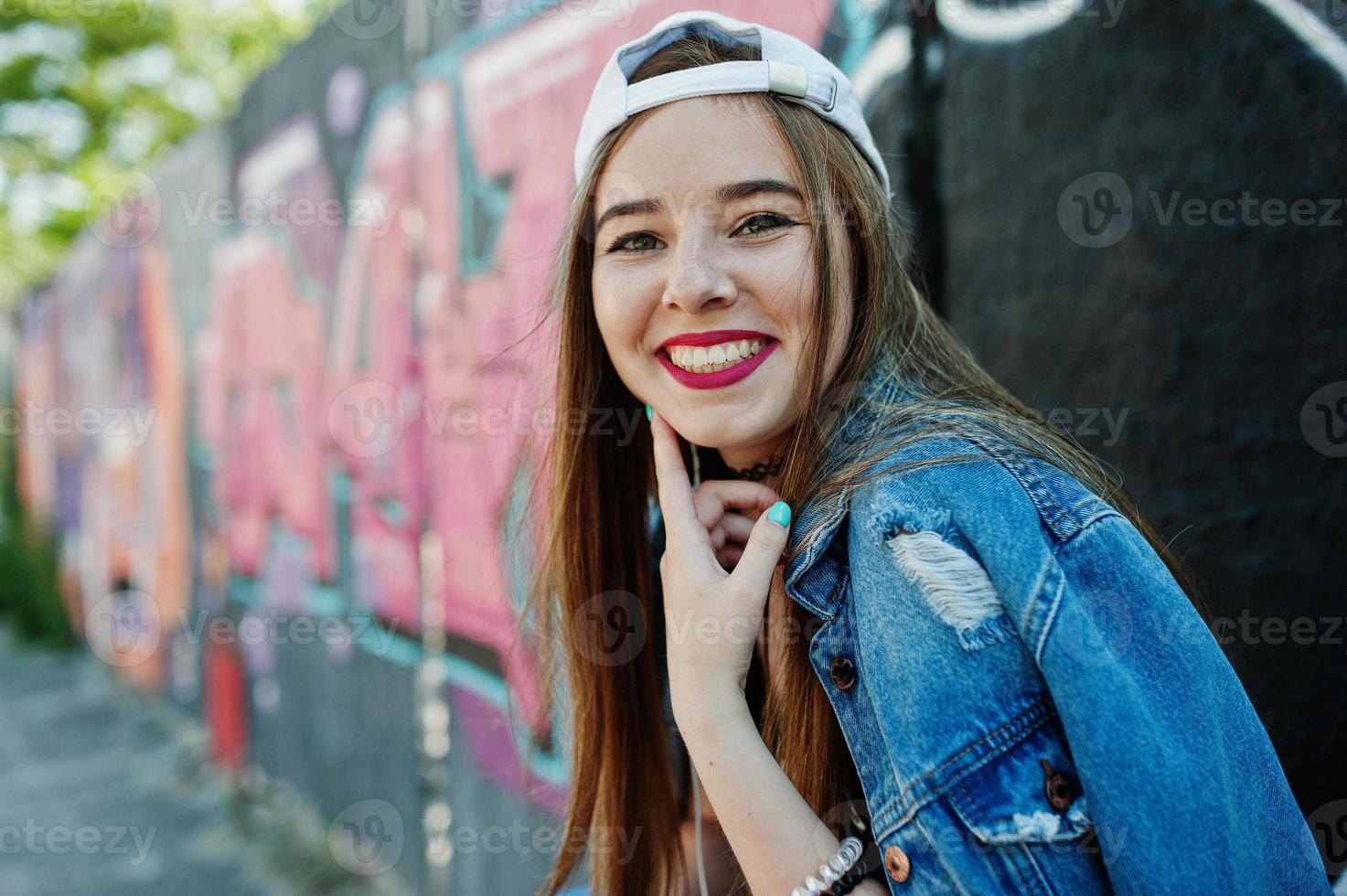 elegante chica hipster casual con gorra y jeans usa música para escuchar desde auriculares de teléfono móvil contra una gran pared de graffiti con bomba. foto