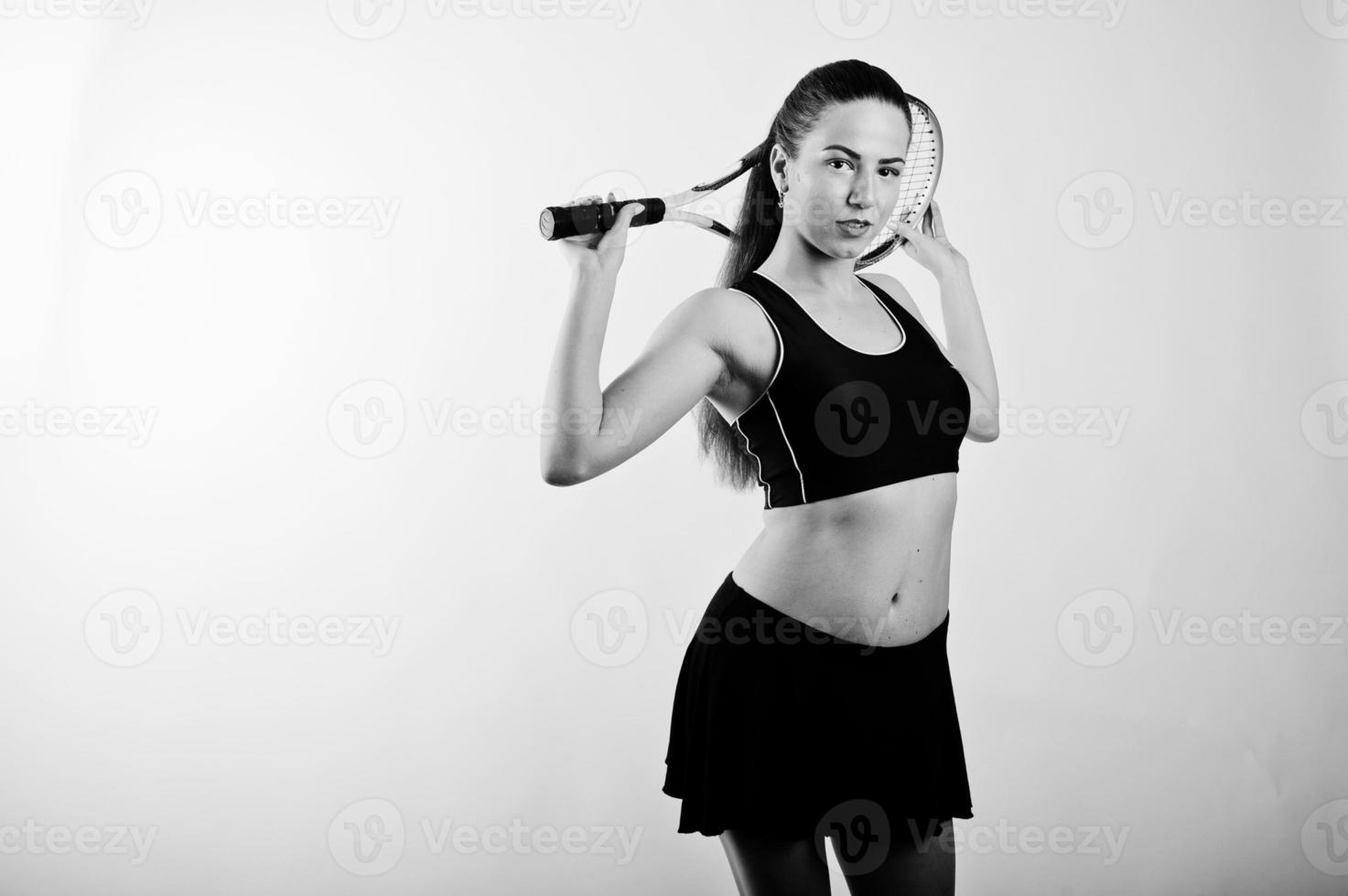 Black and white portrait of beautiful young woman player in sports clothes holding tennis racket while standing against white background. photo