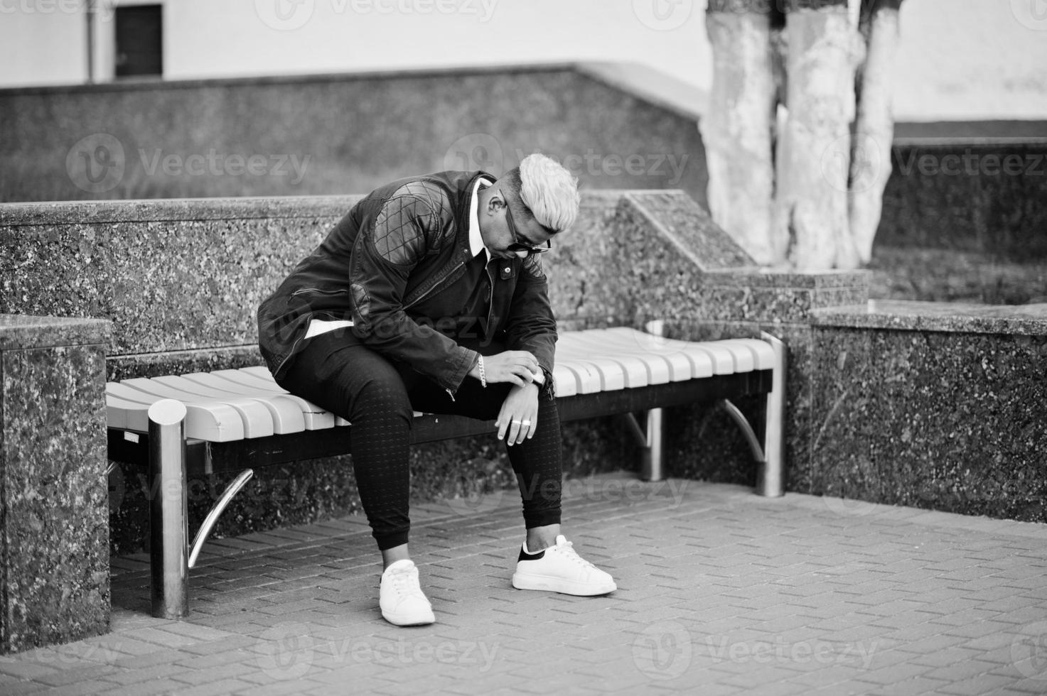 Stylish arabian muslim boy with originally hair and sunglasses posed on streets, sitting on bench. photo