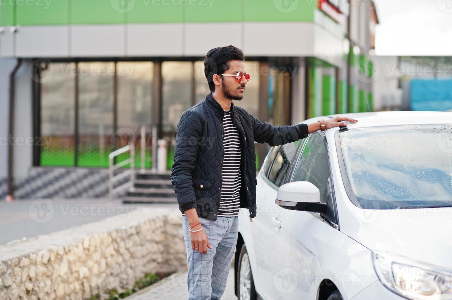 South asian man or indian male wear red eyeglasses stand near his white transportation on car wash. photo