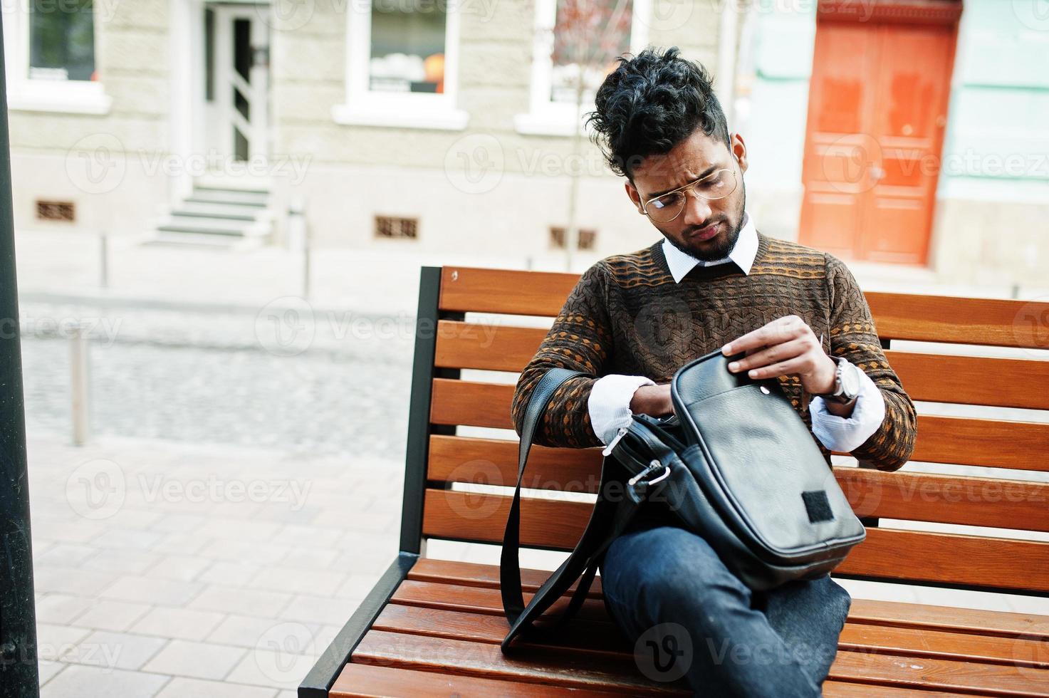 Portrait of young stylish indian man model pose in street, sitting on bench with handbag. photo