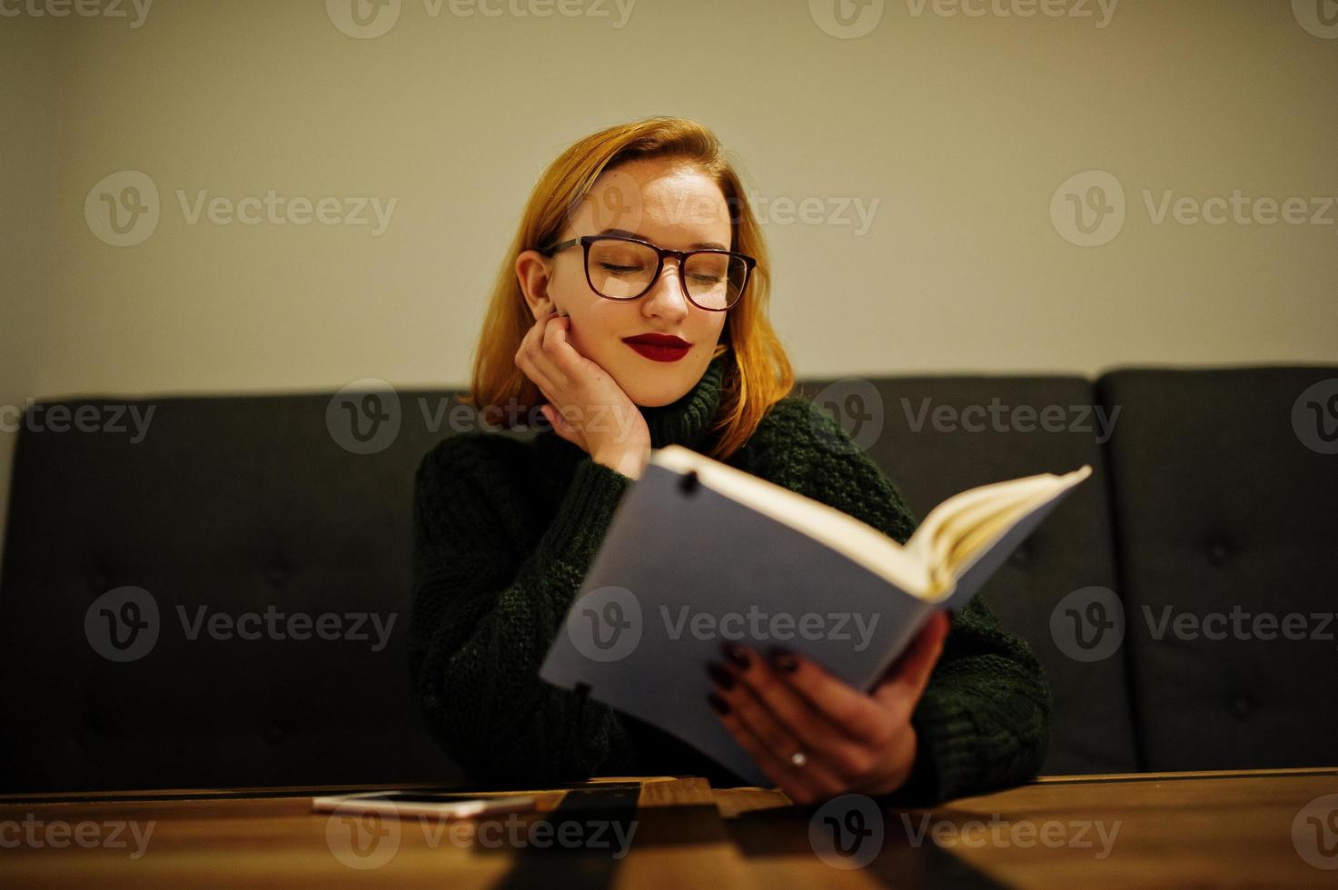 joven alegre y hermosa mujer pelirroja con anteojos, vestida con un suéter verde de lana cálida, sentada en el café y usando su cuaderno. foto