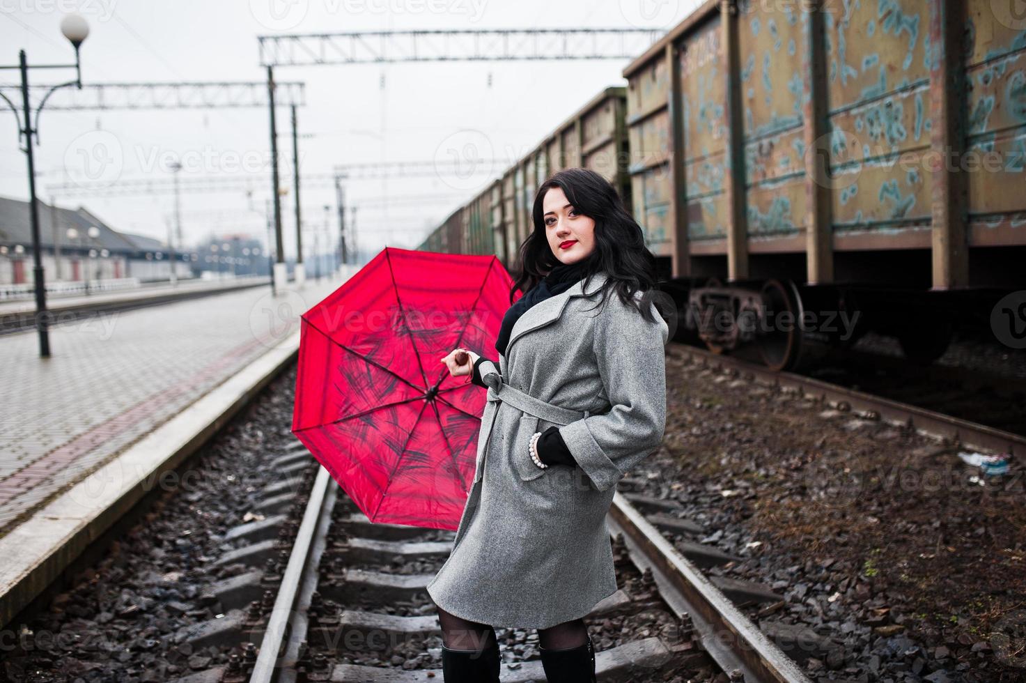Brunette girl in gray coat with red umbrella in railway station. photo
