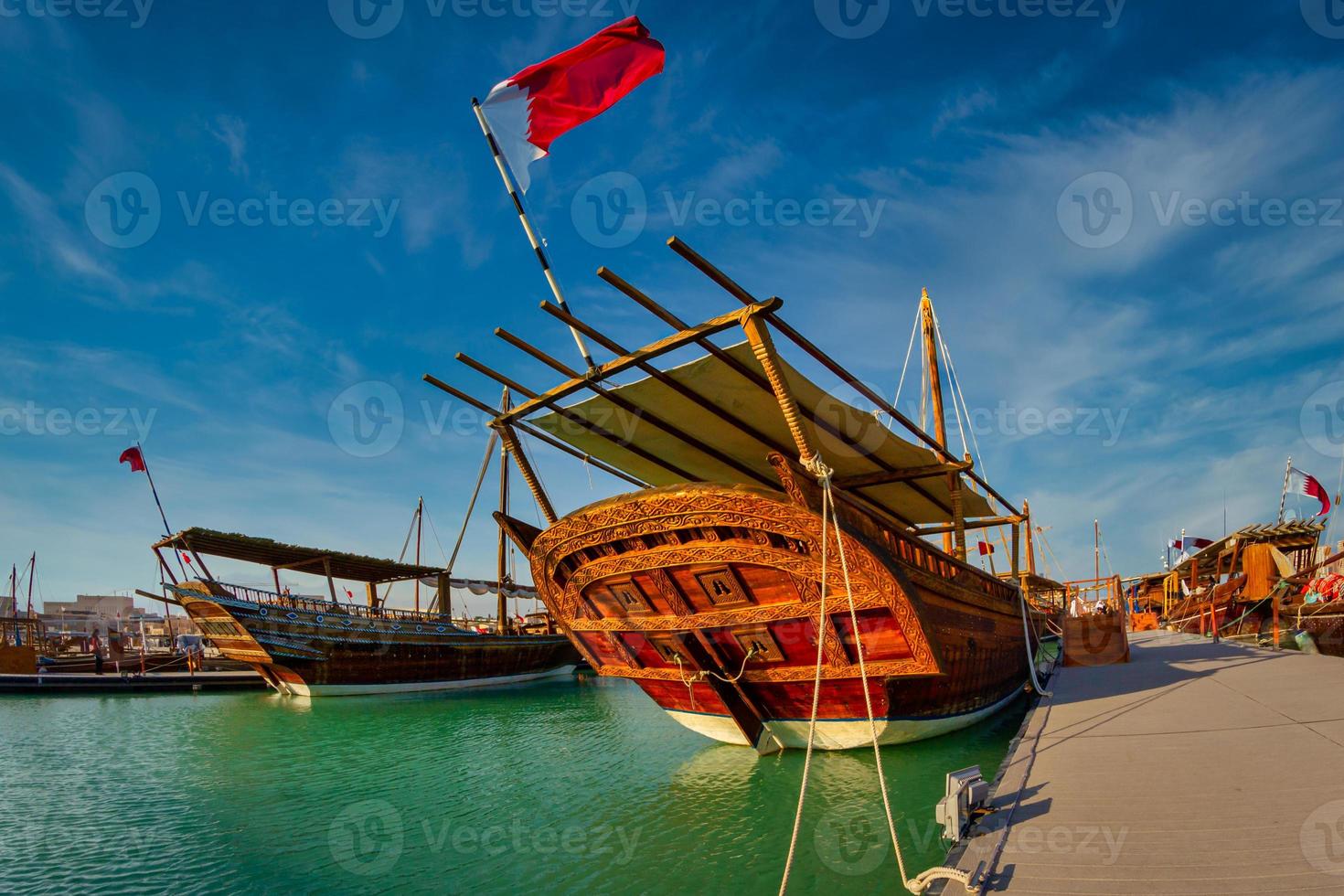 Traditional wooden boats ,dhows, in Katara beach Qatar daylight view with Qatar flag and clouds in sky photo