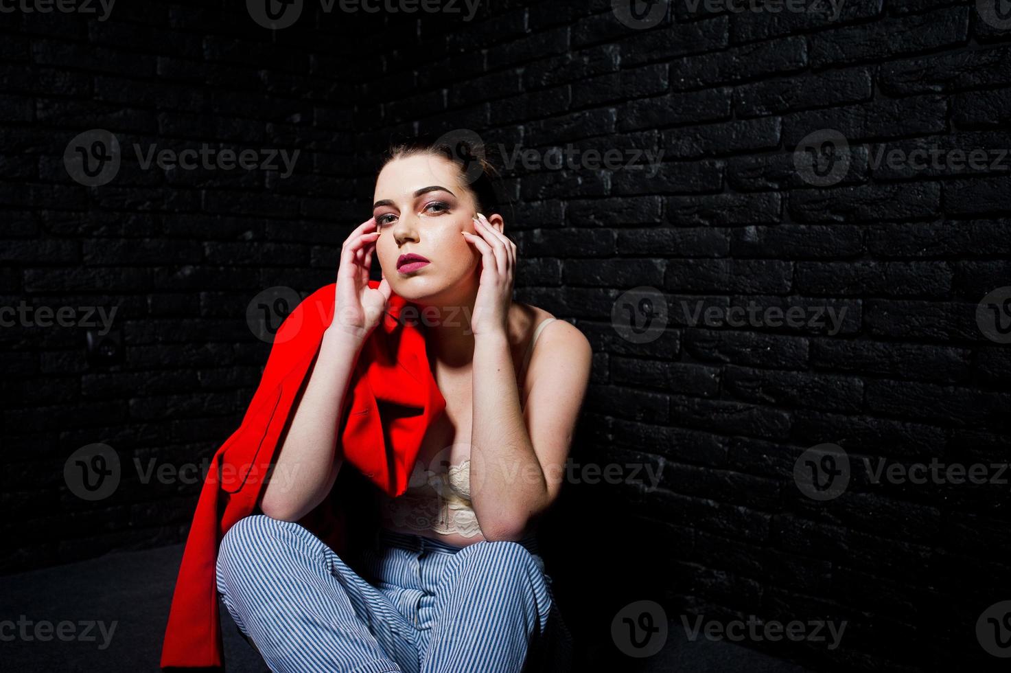 Stylish brunette girl on red jacket against studio black brick wall. photo