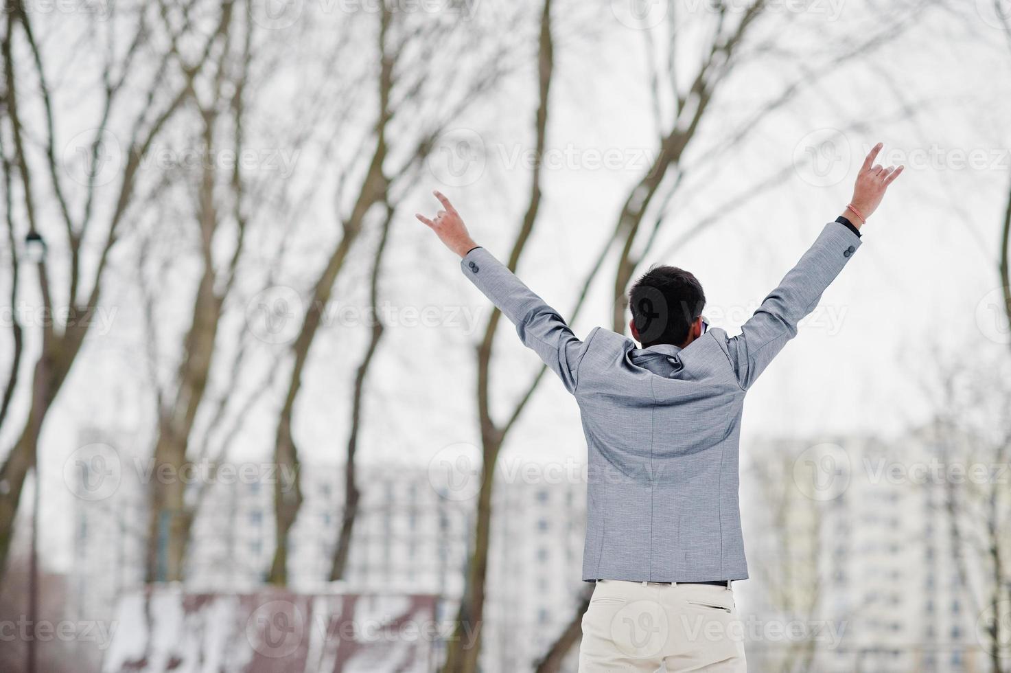 Back of casual young indian man in silver jacket showing rock finger sign, hands up and posed at winter day. photo