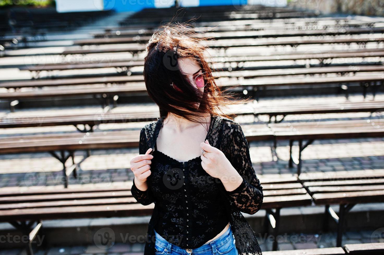 Portrait of brunette girl in pink glasses wear on black posed outdoor on sunny day against row of benches. photo