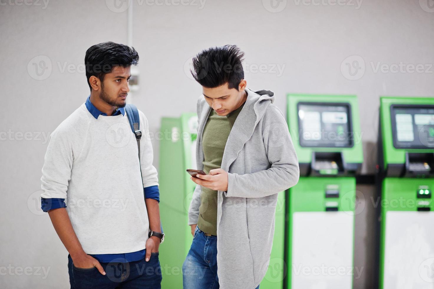 Two asian guys looking at mobile phone against row of green ATM. photo