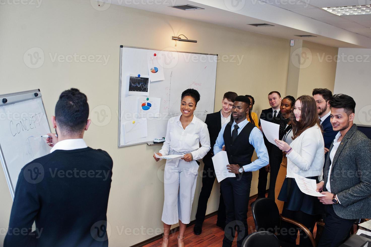 Mixed race business coach presenting report standing near whiteboard pointing on sales statistic shown on diagram and chart teach diverse company members gathered together in conference room. photo