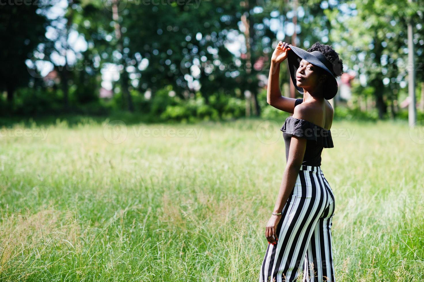 retrato de una hermosa mujer afroamericana de 20 años vestida con pantalones de rayas blancas y negras y sombrero de verano posando en la hierba verde en el parque. foto