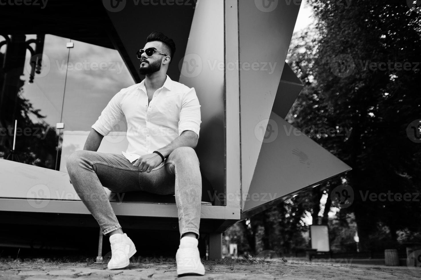 Stylish tall arabian man model in white shirt, jeans and sunglasses sitting at street of city. Beard attractive arab guy. photo