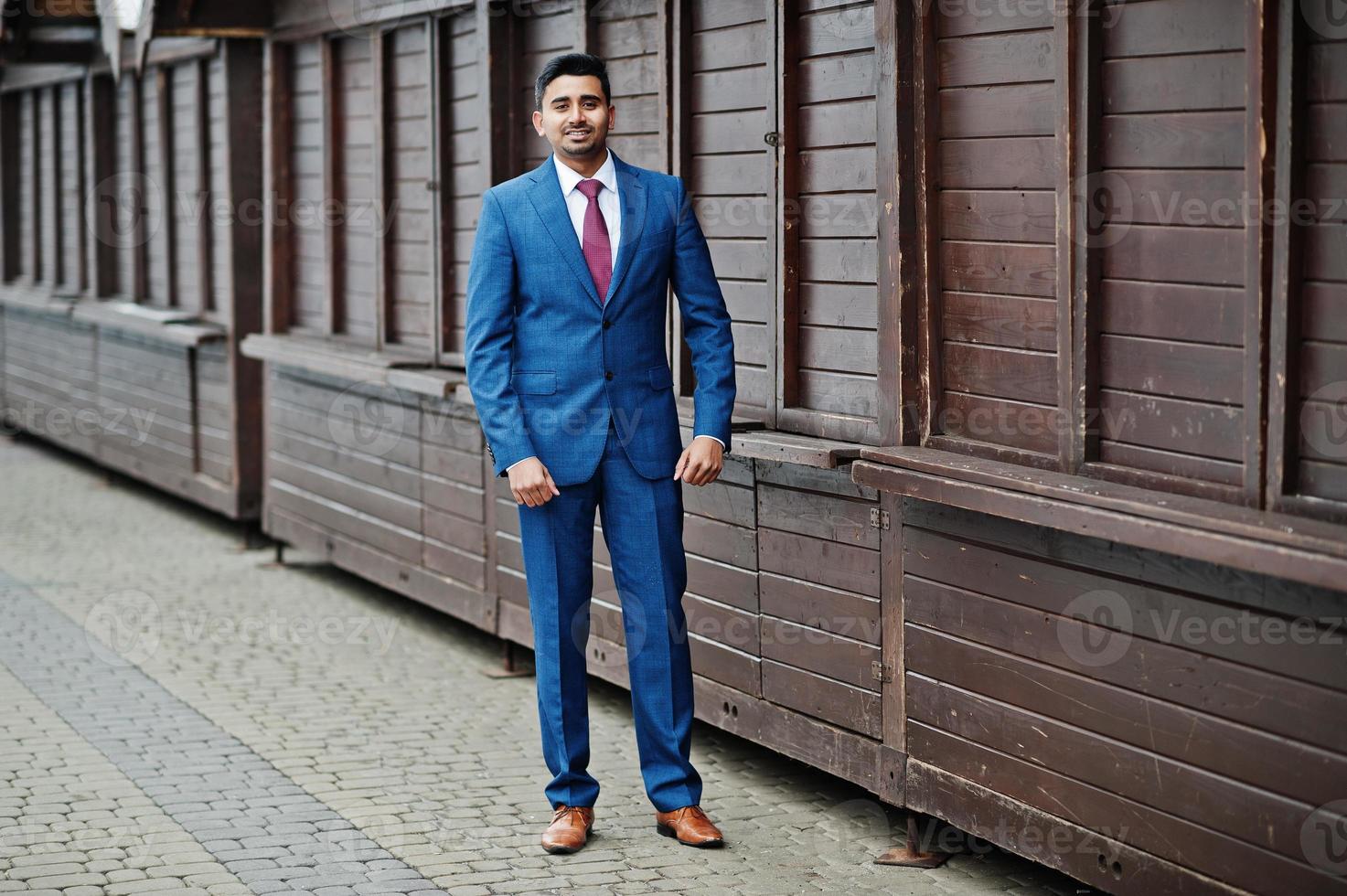 elegante modelo de hombre de moda indio en traje posado contra puestos de madera. foto