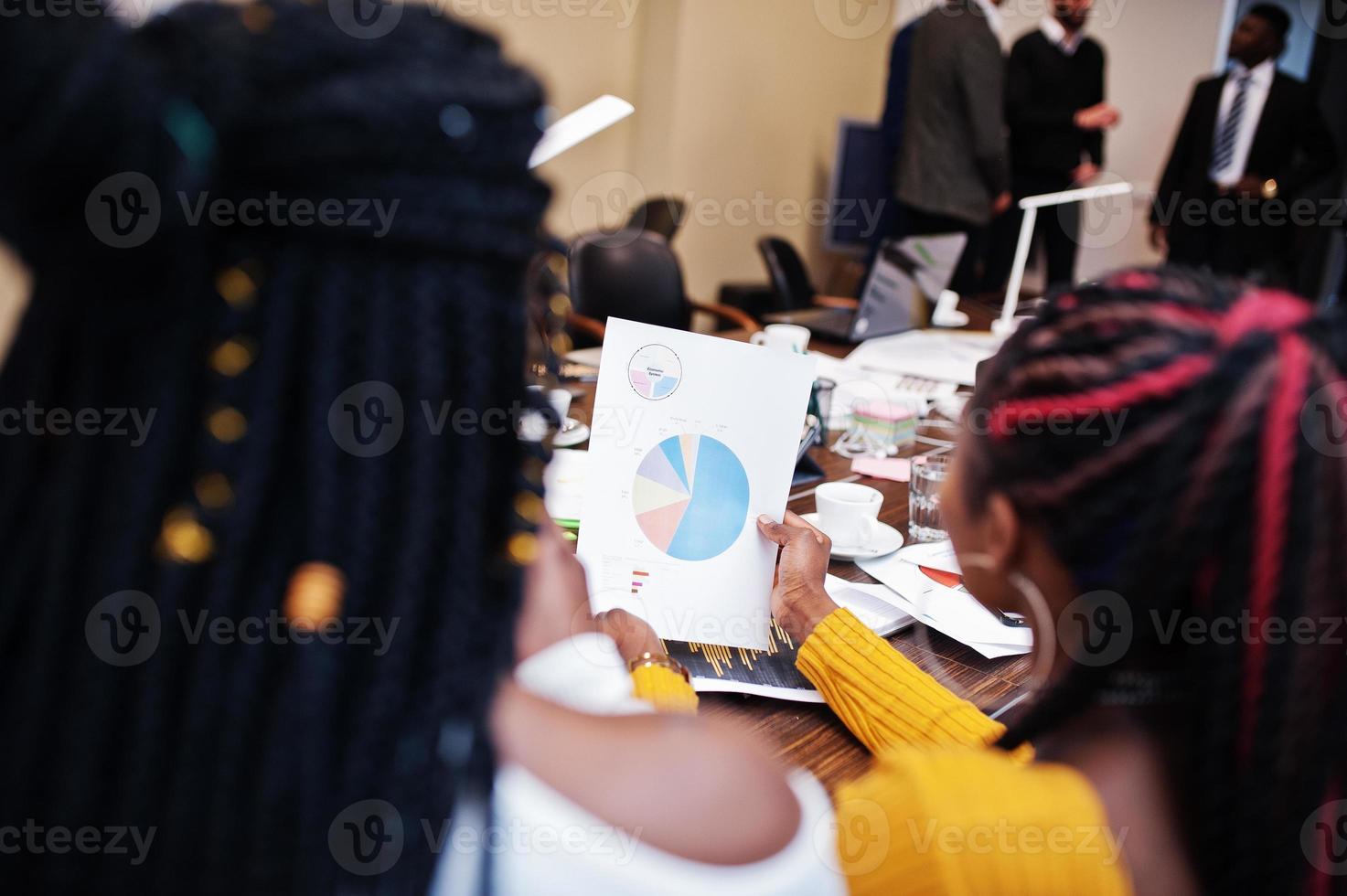 Multiracial women colleagues, crew of divercity female partners in office sit at the table and look at infographic sales schedule on a piece of paper. photo