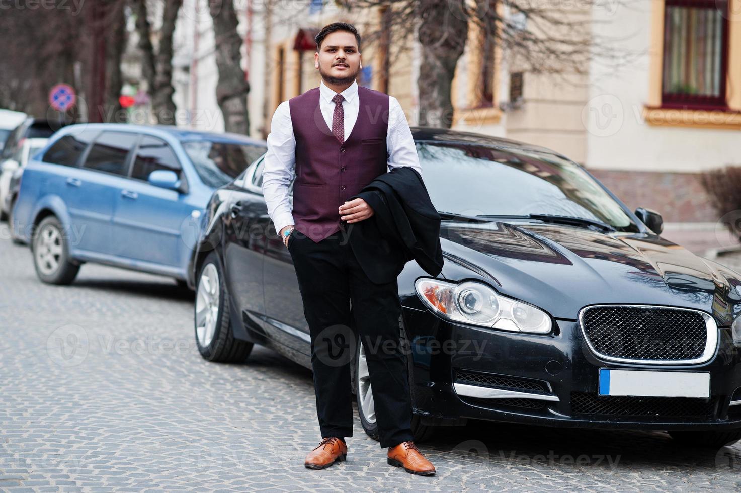 Stylish indian businessman in formal wear vest suit standing against black business car on street of city. photo