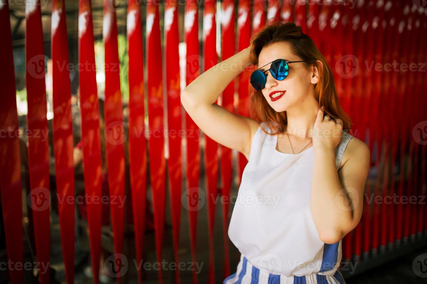 Attractive redhaired woman in sunglasses posing against red background. photo