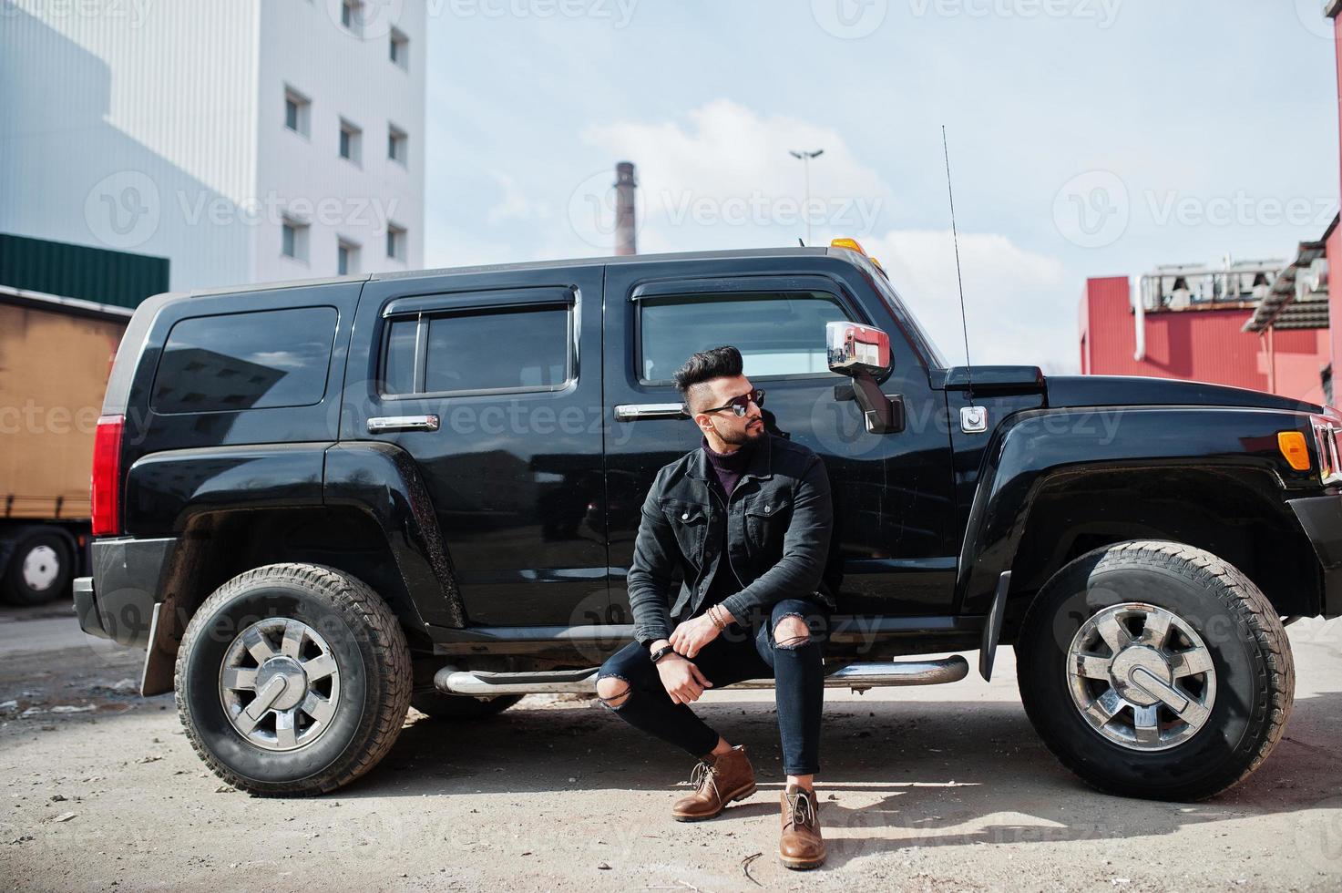Fashion rich beard Arab man wear on black jeans jacket and sunglasses posed against big black suv car. Stylish, succesful and fashionable arabian model guy. photo
