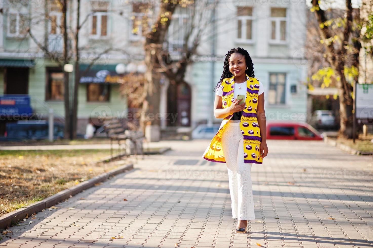Stylish african american women in yellow jacket posed on street at sunny day with mobile phone at hand. photo