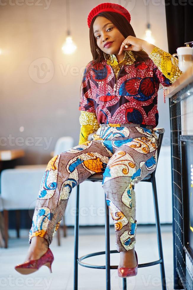 Enthusiastic african american woman in trendy coloured outfit with red beret chilling in cozy cafe, sitting on bar counter. photo