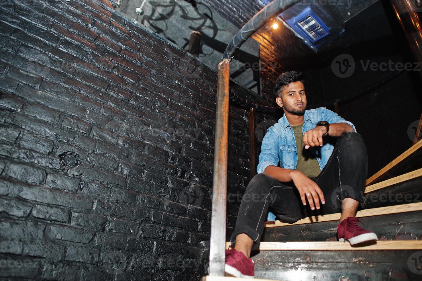 Portrait of handsome successful bearded south asian, young indian freelancer in blue jeans shirt standing in night club on stairs. photo