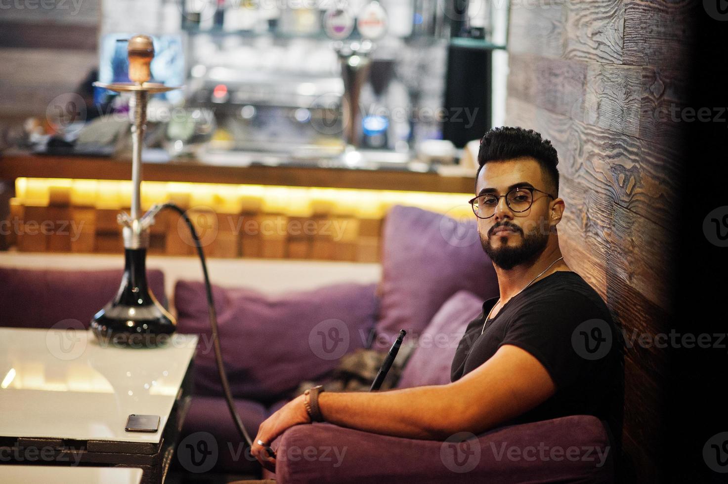 Stylish beard arabian man in glasses and black t-shirt smoking hookah indoor bar. Arab model having rest. photo