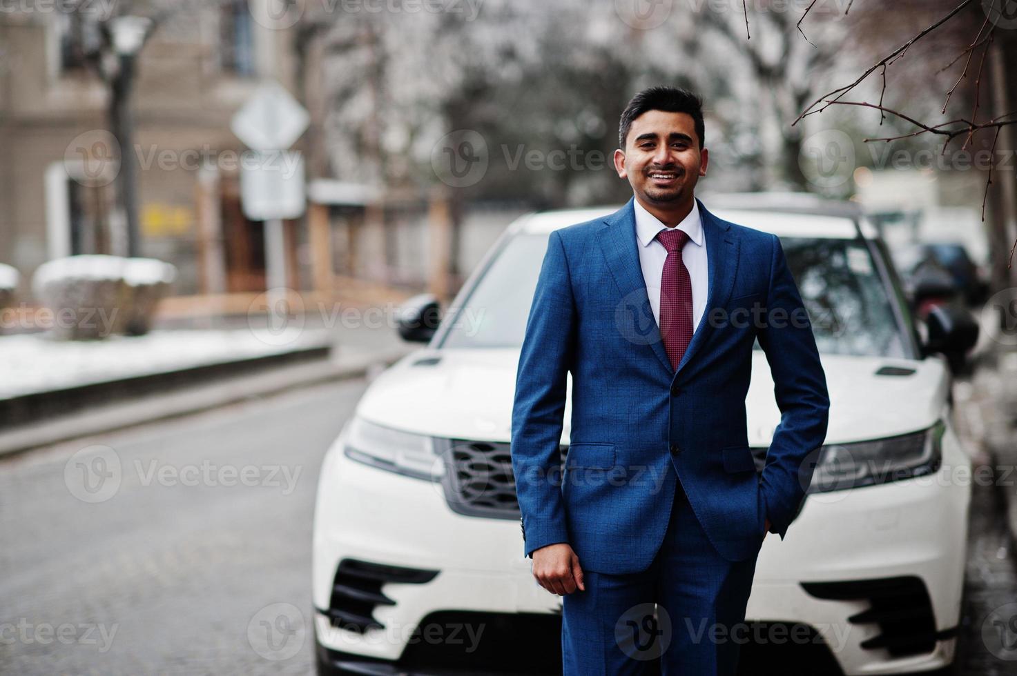Elegant indian fashionable man model on suit posed at winter day against white business suv car. photo
