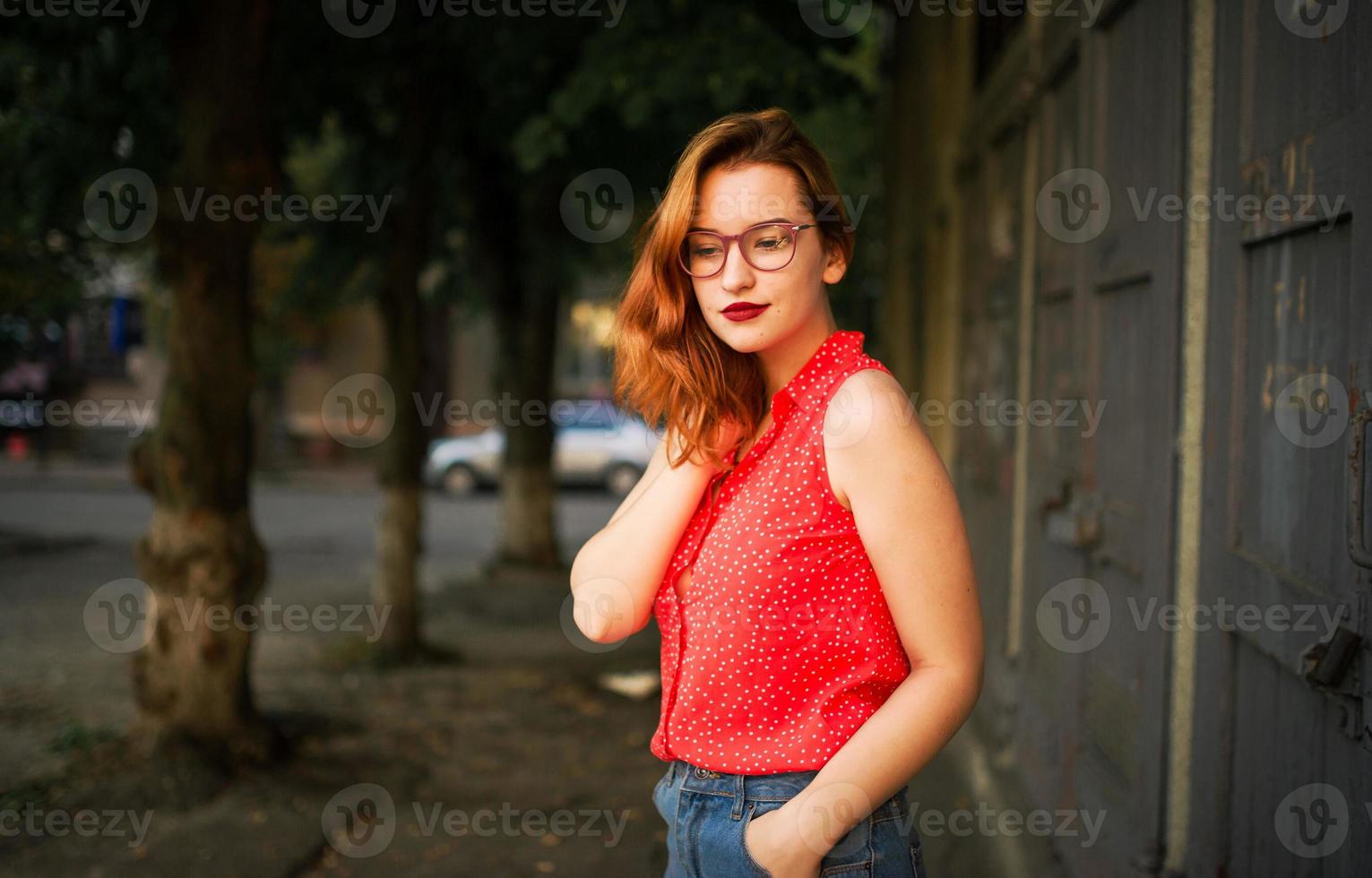 Attractive redhaired woman in eyeglasses, wear on red blouse and jeans skirt posing. photo